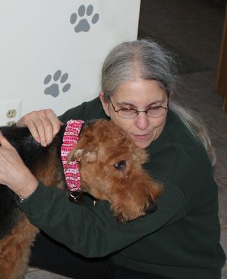 A woman with glasses gently hugs a brown airedale terrier next to a white wall with paw prints.