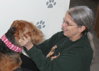 An older woman smiles as she pets a large dog with a red bandana in a room adorned with paw prints on the wall.