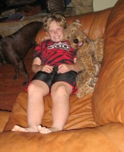 A young boy smiling, sitting on a brown leather couch with a brown poodle, with another darker dog in the background.