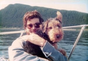 Man with sunglasses holding a terrier dog on a boat, with a forested shoreline and clear sky in the background.