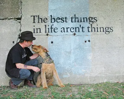 A man in a hat sitting beside a dog, both facing a wall with the phrase "the best things in life aren't things" painted on it.