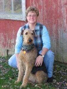 A woman in glasses and a blue shirt sits beside a large, brown dog in front of a red barn door.