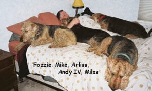 Man sleeping in bed with five large dogs, each labeled with names: fozzie, mike, arliss, andy iv, miles.