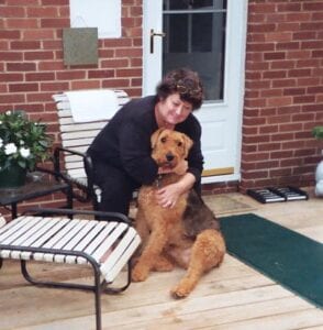 A woman in a black outfit cuddling a large brown dog on a porch next to potted plants and a white bench.