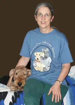 A woman in a blue t-shirt featuring cartoon dogs sits with a brown terrier on her lap, against a brown background.