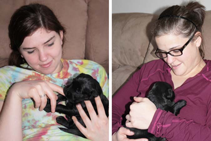 Two women holding black puppies, sitting on a couch; one woman is wearing a tie-dye shirt, the other in a maroon top.