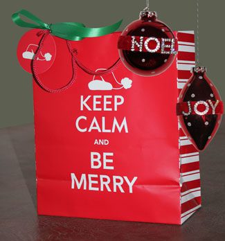 A bright red holiday gift bag with the text "keep calm and be merry" on it, adorned with a green ribbon and two red christmas ornaments reading "noel" and "joy".