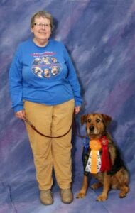 A senior woman in a blue sweater stands next to a dog with ribbons around its neck, both posing against a purple backdrop.