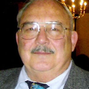 Elderly man with glasses and a mustache, wearing a light gray suit and a patterned tie, smiling slightly in a dimly lit room.
