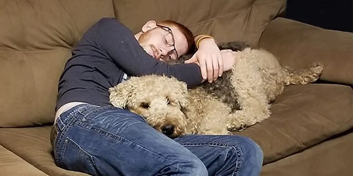 A man with glasses sleeps on a couch, embracing a large, fluffy dog.