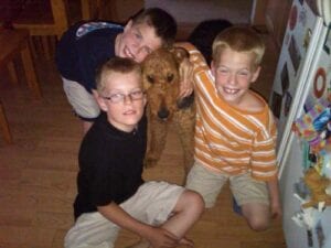 Three boys smiling with a brown dog in a home setting.
