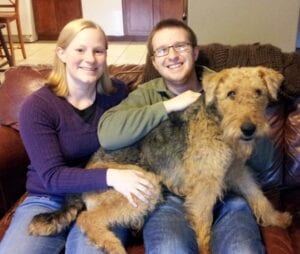 A smiling couple seated on a couch with a large airedale terrier dog in their lap.