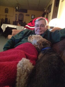 Elderly man wearing glasses and a santa hat relaxing on a couch with two dogs on his lap in a cozy room.