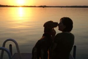 A woman and a dog sitting on a dock, facing a sunset over a lake, with the woman kissing the dog on the nose.