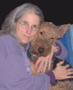 An elderly woman with glasses embracing a brown airedale terrier dog, both looking towards the camera.
