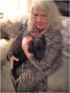 Blurred image of a smiling woman with white hair, wearing glasses and a patterned top, holding a small black dog.