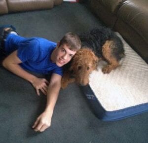 Young man in blue shirt smiling next to a large brown dog on a white dog bed in a living room.