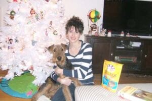 A woman sitting with a dog beside a decorated christmas tree in a living room.