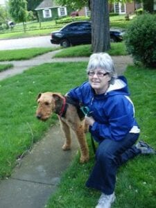 A woman kneeling beside a large brown dog on a leash, both posing for the photo on a sidewalk in a residential area.