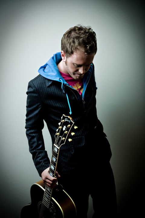 Man in a suit and tie with an unbuttoned shirt, holding an acoustic guitar, looking thoughtfully downwards.