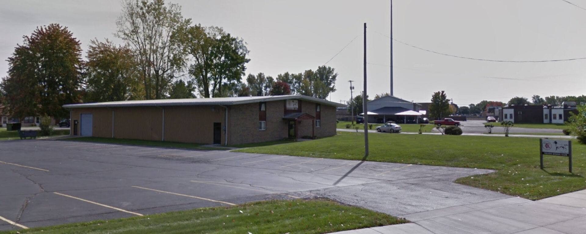 A single-story brick building with a large parking lot in a rural setting, surrounded by a few sparse trees and utility poles under a clear sky.